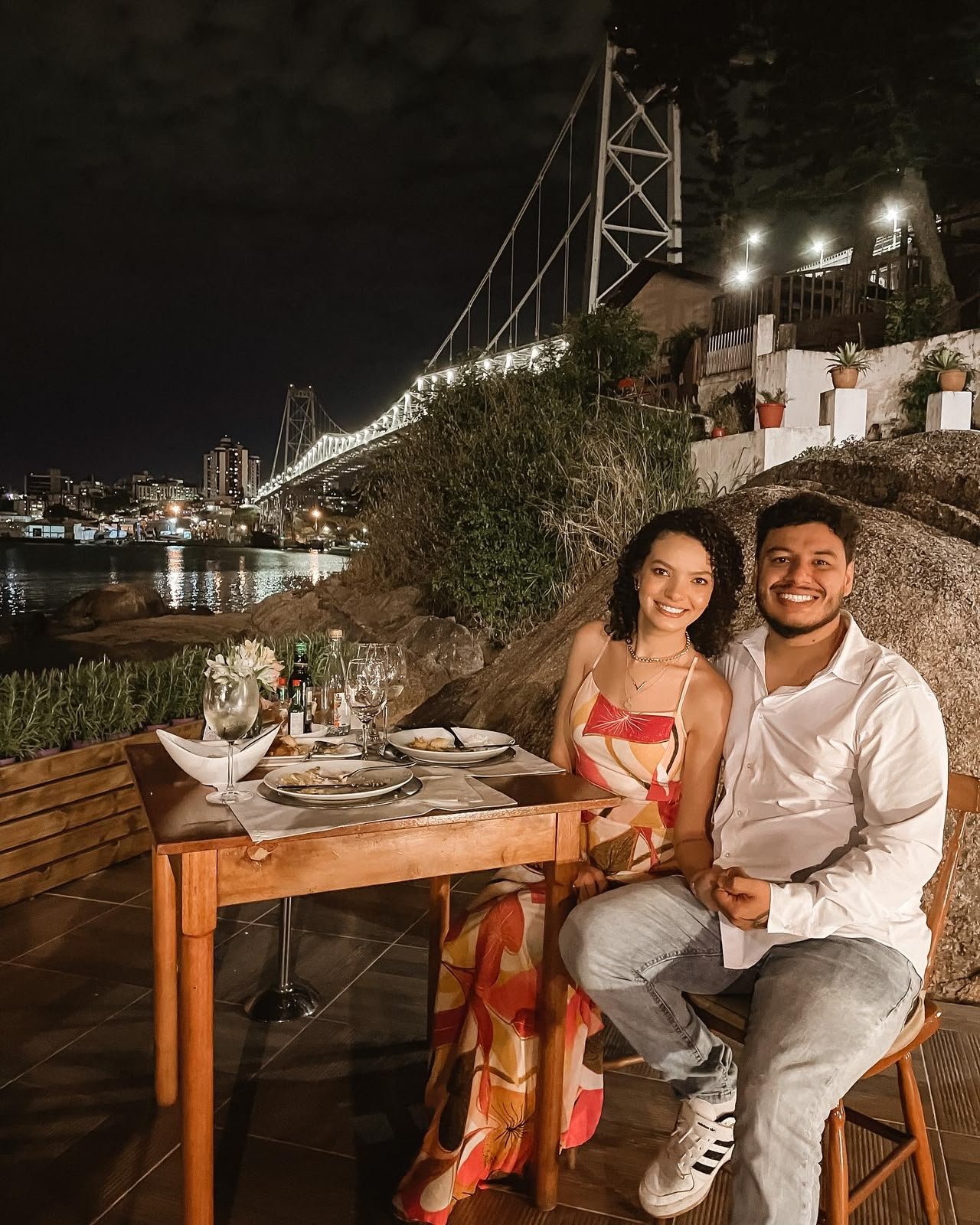 Cássia e Pablo sorrindo em jantar à beira da ponte, à noite