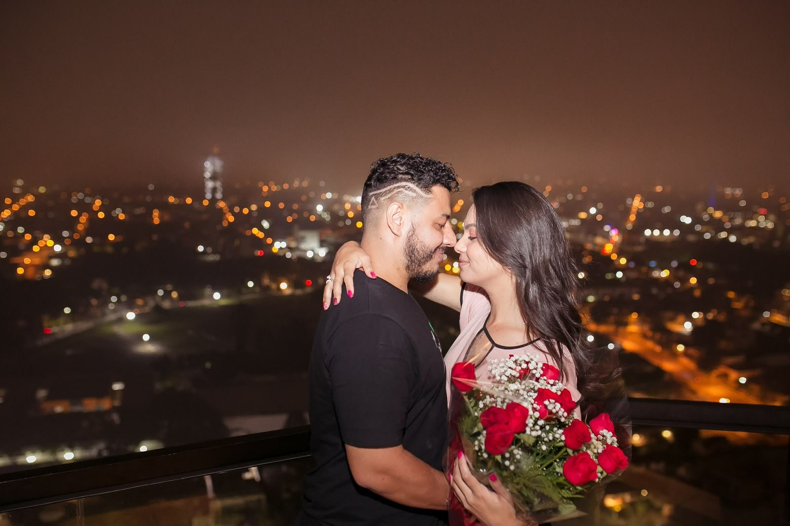 Cássia e Pablo em um terraço à noite, com buquê de rosas